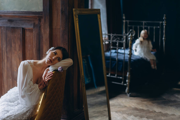 mulher em um belo vestido de tule branco sentado em uma cadeira e silhueta borrada da menina em um vestido branco boêmio sentado na cama atrás do espelho  - Foto, Imagem