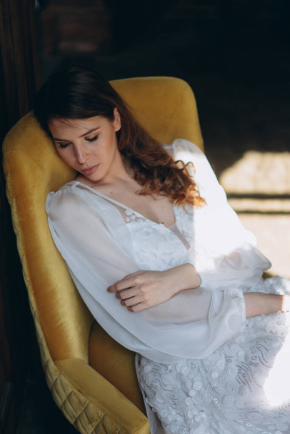  girl in a beautiful white bohemian tulle dress sitting in a chair near the balcony, closeup view of woman in a pensive mood and she looking down - Foto, imagen