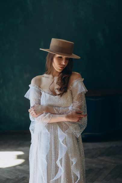  girl in a beautiful white sensual tulle dress and in hat standing in the blue room and posing for the camera - Foto, immagini