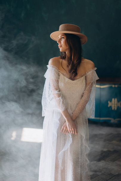  girl in a beautiful white sensual tulle dress and in hat standing in the blue room and posing for the camera - Foto, imagen