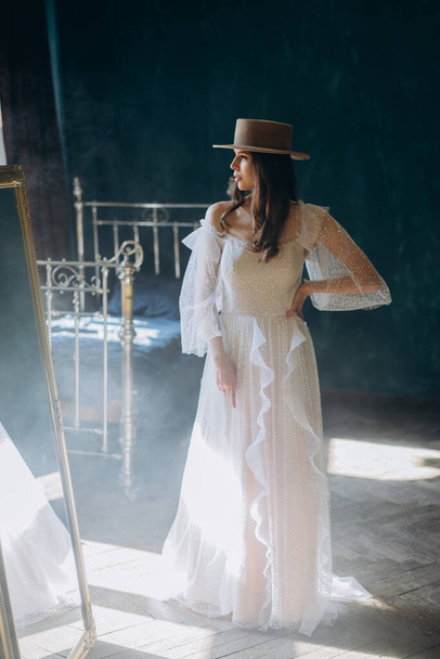  girl in a beautiful white sensual tulle dress and in hat standing in the blue room and posing for the camera - Foto, Imagen