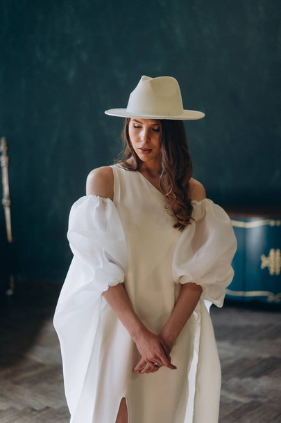 girl in a beautiful white sensual dress and white hat standing in the blue room and posing for the camera - Foto, afbeelding