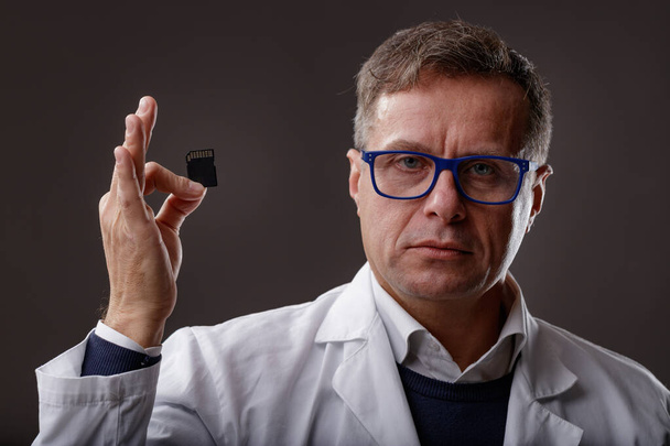 Ekspert naukowy lub śledczy w białym fartuchu laboratoryjnym trzymający kartę SD z cyfrowymi danymi w ręku podczas dochodzenia na ciemnym tle - Zdjęcie, obraz