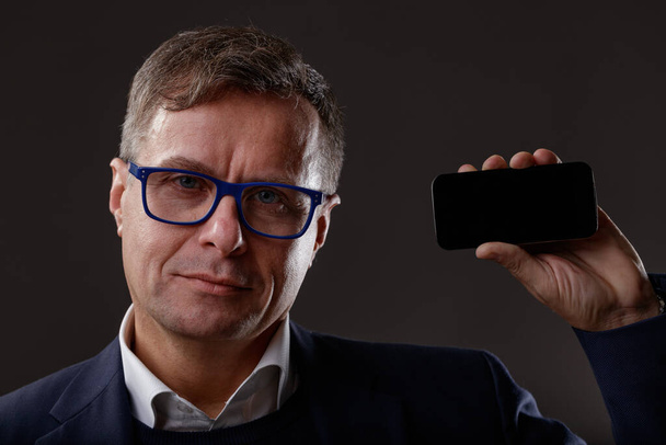 Studio portrait of a middle-aged man looking at camera with confidence while holding and showing his performant smartphone useful for online communication and information - Photo, image