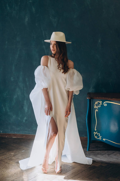 girl in a beautiful white sensual dress and white hat standing in the blue room and posing for the camera - Photo, Image