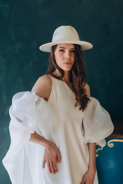 girl in a beautiful white sensual dress and white hat standing in the blue room and posing for the camera - Foto, imagen