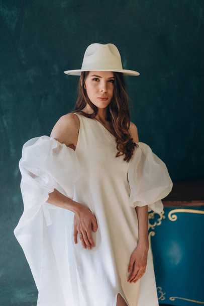girl in a beautiful white sensual dress and white hat standing in the blue room and posing for the camera - Foto, Imagem