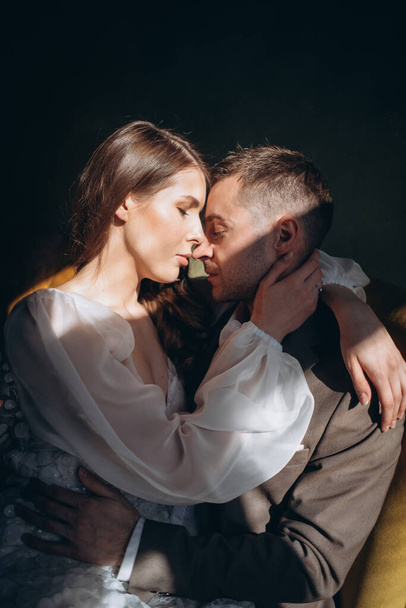 man in the elegant suit sitting in the chair and a girl in a beautiful white sensual tulle dress kissing him by the window in warm light - Foto, Imagem