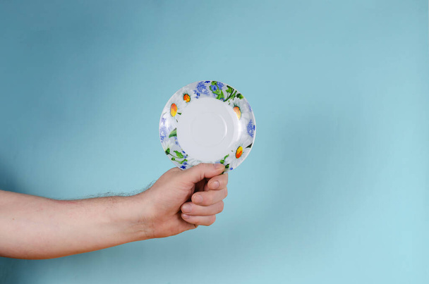 Mężczyzna trzymający biały spodek z pomalowanymi kwiatami. Ceramiczny spodek jasno zdobiony w kolorach niebieskim i żółtym. Białe stokrotki na naczyniach. - Zdjęcie, obraz