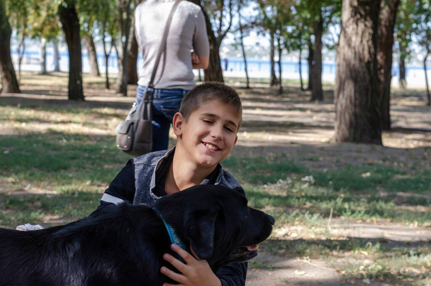 Συναισθηματικό πορτραίτο ενός αγοριού να χαϊδεύει ένα μαύρο σκύλο με ένα μπλε κολάρο στο πάρκο. Ένα 10χρονο παιδί και ένα 11χρονο θηλυκό Λαμπραντόρ. Μια βόλτα στο πάρκο με ένα κατοικίδιο. - Φωτογραφία, εικόνα