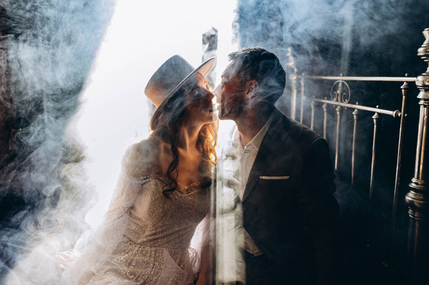 man in elegant suit and girl in a beautiful white sensual tulle dress and hat sitting on bed and kissing in the smoky room - Fotó, kép