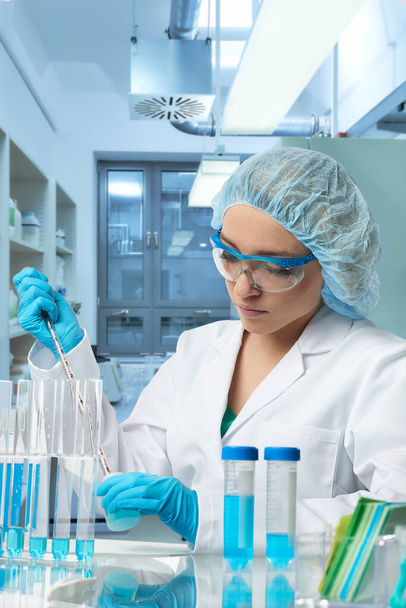 Pharma female tech works in laboratory. Caucasian, european woman, young adult in protective gloves, hat and white gown handles sample tube. Defocused lab interior, selective focus. Biotech research. - Foto, Imagen