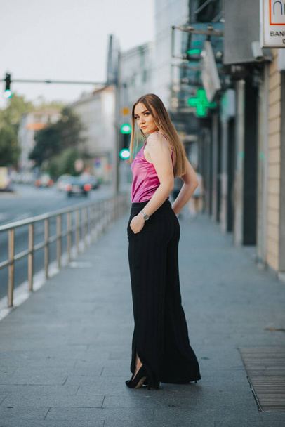 A vertical shot of a young blonde female model posing on the sidewalk in a pink blouse and black pants - Фото, изображение