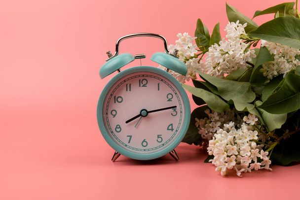 Синий винтажный будильник с белыми сиреневыми цветами, изолированный на розовом фоне. Смена весеннего времени, весенние цветы и будильник.  - Фото, изображение