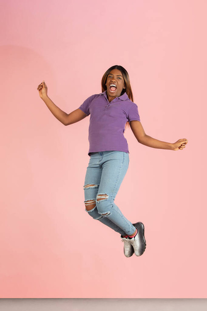 Ένα χαρούμενο κορίτσι από την Αφρική να πηδάει απομονωμένο σε ροζ φόντο στούντιο. - Φωτογραφία, εικόνα