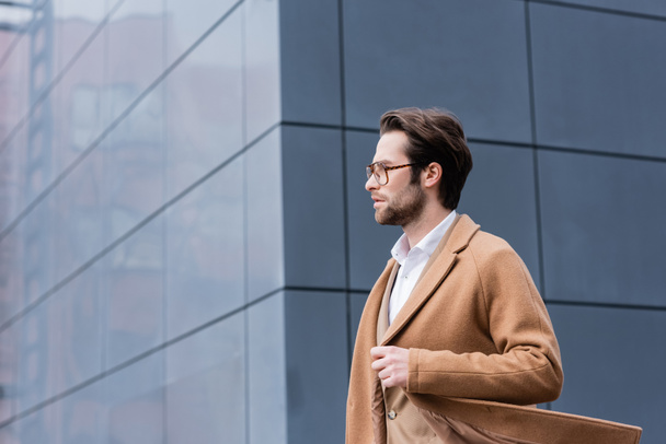 side view of bearded businessman in glasses and beige coat walking outside - Фото, изображение