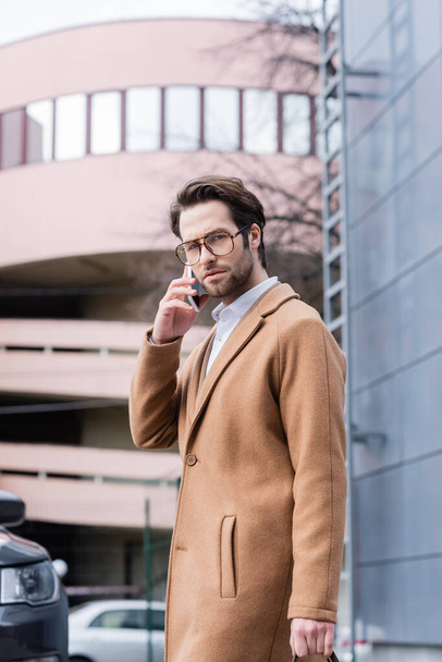 young businessman in glasses talking on smartphone near building  - Fotó, kép