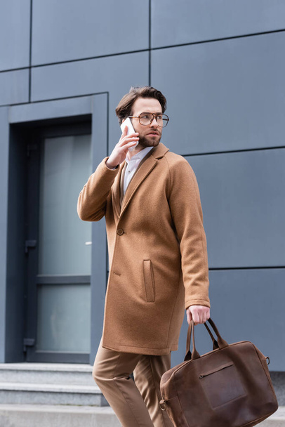 fiatal férfi szemüveg és kabát beszél okostelefon közelében épület  - Fotó, kép