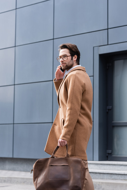 young man in glasses and coat talking on cellphone near building  - Фото, зображення