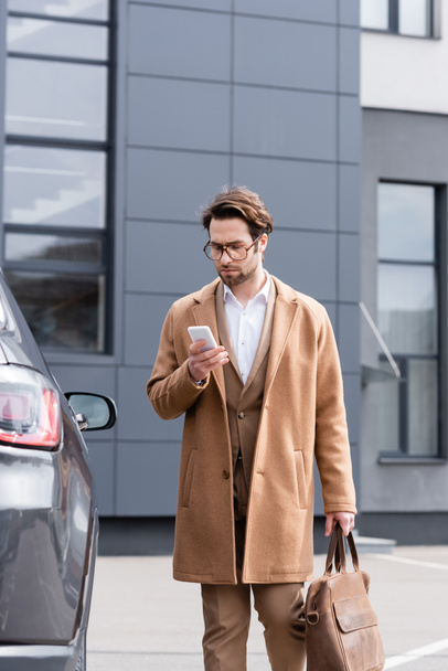 businessman in glasses and coat texting on smartphone near car  - Zdjęcie, obraz