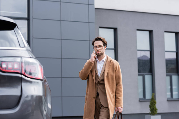 confident businessman in glasses and beige coat talking on mobile phone near car - Foto, afbeelding