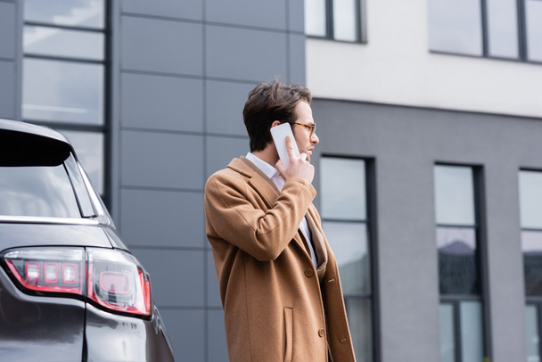 side view of confident man in glasses and beige coat talking on mobile phone near car - Фото, изображение