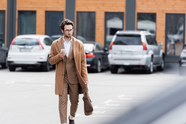 confident businessman in glasses and beige coat walking with leather bag and mobile phone on parking outside - Fotografie, Obrázek