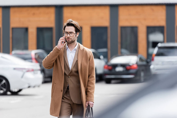 hombre confiado en gafas y abrigo beige hablando en el teléfono móvil en el aparcamiento al aire libre  - Foto, Imagen