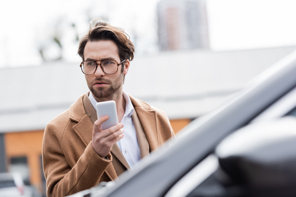 confident man in glasses and beige coat holding mobile phone near blurred car - Foto, immagini