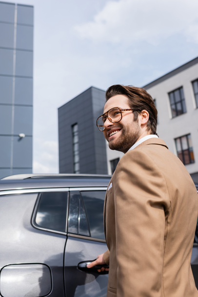 heureux jeune homme d'affaires en veste beige souriant près de la voiture moderne  - Photo, image
