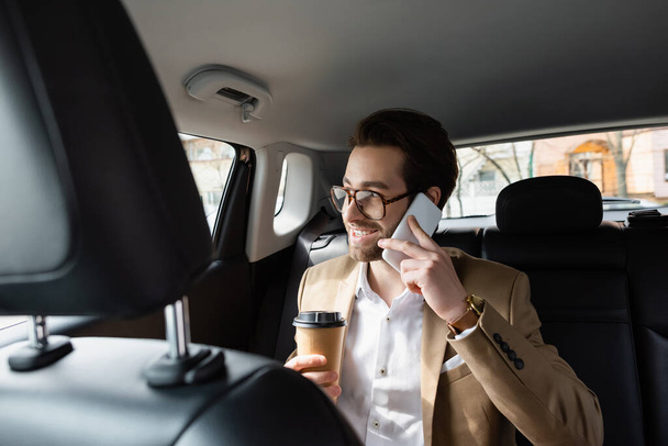 hombre feliz con traje y gafas sosteniendo la taza de papel y el teléfono inteligente hablando en el coche  - Foto, Imagen