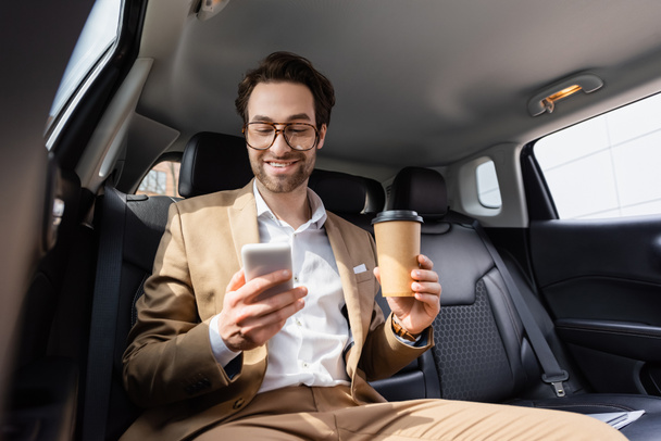joyful businessman in suit and glasses holding paper cup and smartphone in car  - 写真・画像