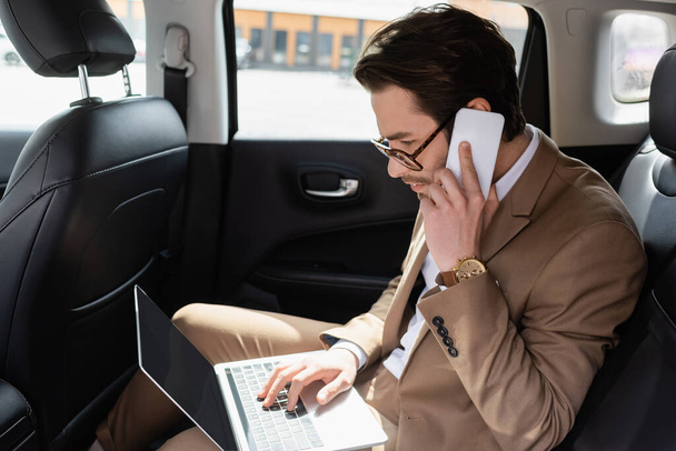 businessman in glasses working remotely with laptop while talking on smartphone in car - Photo, Image