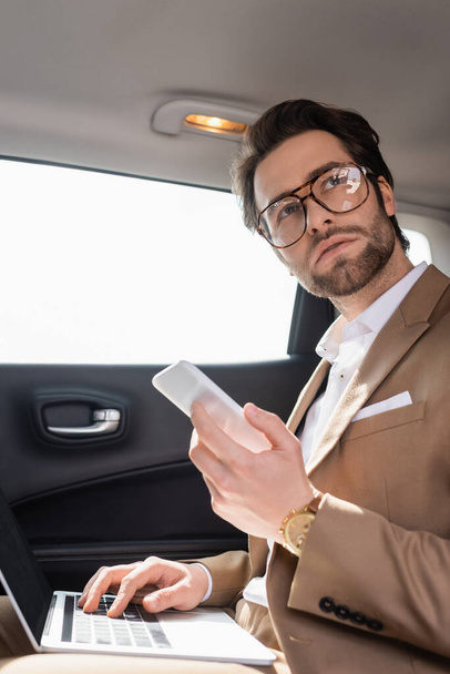 businessman in glasses working remotely with laptop while holding smartphone in car - 写真・画像