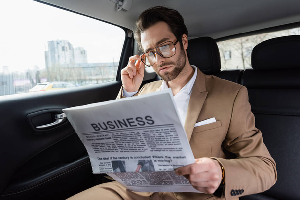 confident man adjusting glasses and reading business newspaper in car  - Fotó, kép