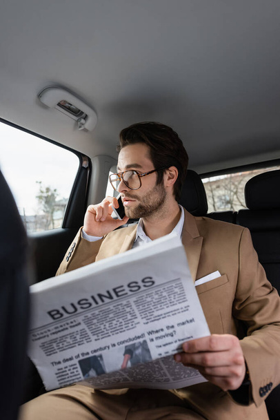 уверенный в себе человек в очках разговаривает на смартфоне и держит деловую газету в машине  - Фото, изображение
