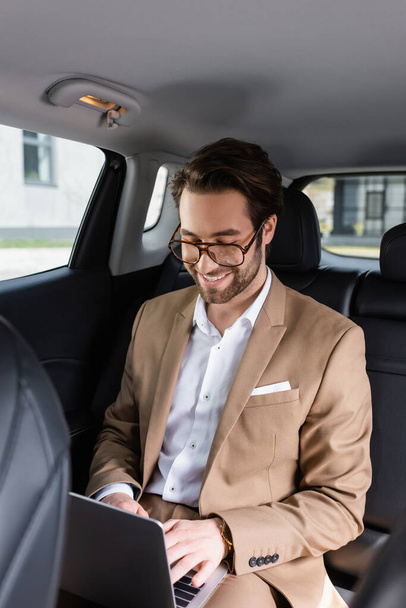 cheerful businessman in glasses typing on laptop in car - Foto, Bild