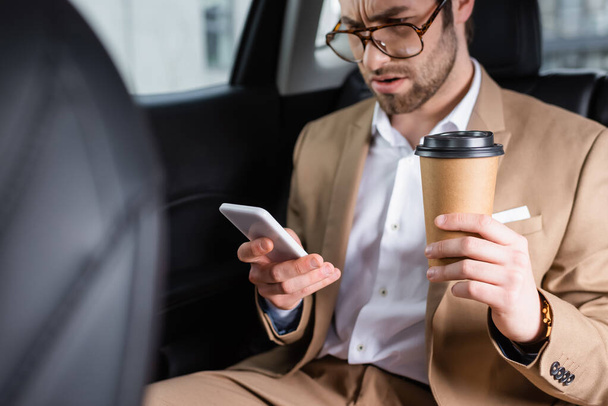 displeased man in suit and glasses holding paper cup and looking at smartphone in car  - Photo, Image