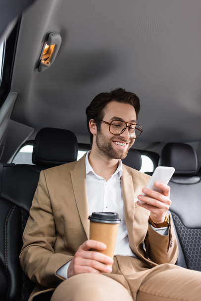 happy businessman in suit and glasses holding paper cup and smartphone in car  - Foto, afbeelding