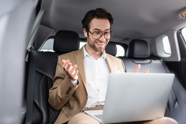 smiling businessman in glasses and wireless earphones gesturing during video call in car  - Foto, Bild