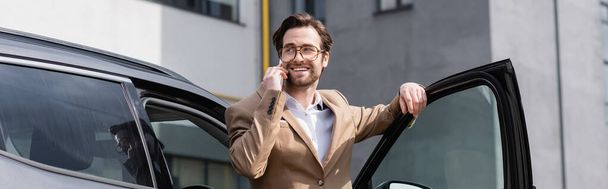 glücklicher Mann in Jacke und Brille, der mit Smartphone spricht und neben Auto mit geöffneter Tür steht, Banner - Foto, Bild
