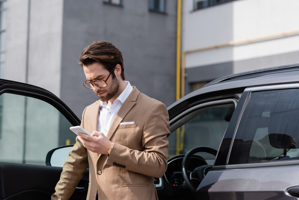 Bärtiger Mann in Anzug und Brille schaut auf Handy und steht neben Auto mit geöffneter Tür - Foto, Bild
