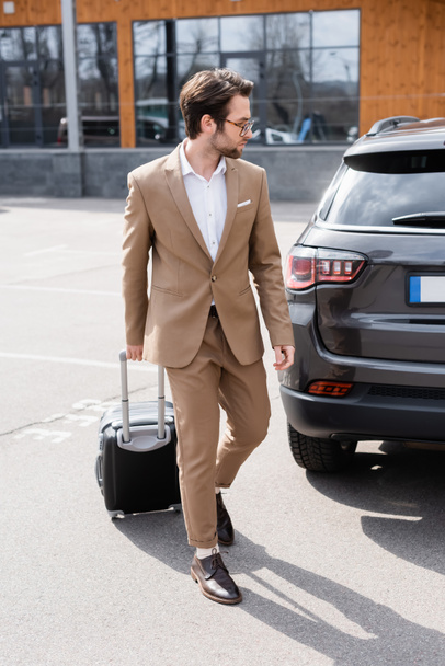 full length of bearded man in suit and glasses walking with suitcase near car on parking  - 写真・画像