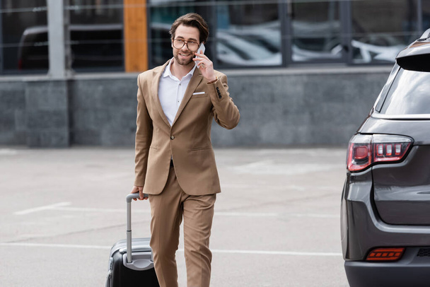 cheerful man in suit and glasses talking on smartphone while walking near car with suitcase - Photo, Image
