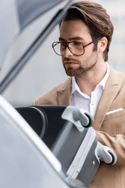 bearded man in glasses and suit putting suitcase in blurred car trunk  - Foto, Imagem