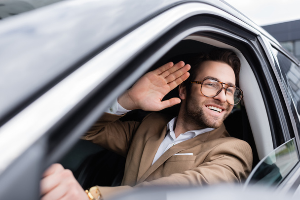 happy man in glasses looking through car window and waving hand while driving auto  - Fotó, kép