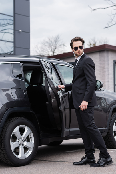 bearded bodyguard in suit and sunglasses with security earpiece opening car door  - Photo, Image