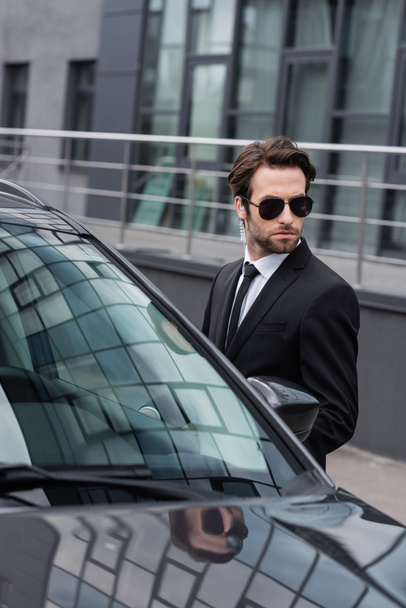 bearded bodyguard in suit and sunglasses with security earpiece near modern auto  - Foto, afbeelding