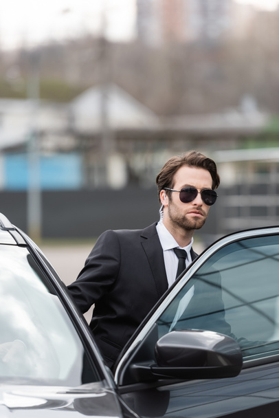bearded bodyguard in suit and sunglasses with security earpiece sitting in modern car  - Zdjęcie, obraz