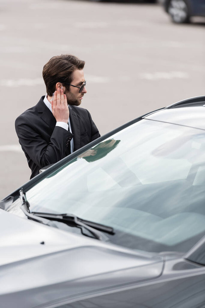 safeguard in suit and sunglasses adjusting security earpiece near car - Zdjęcie, obraz